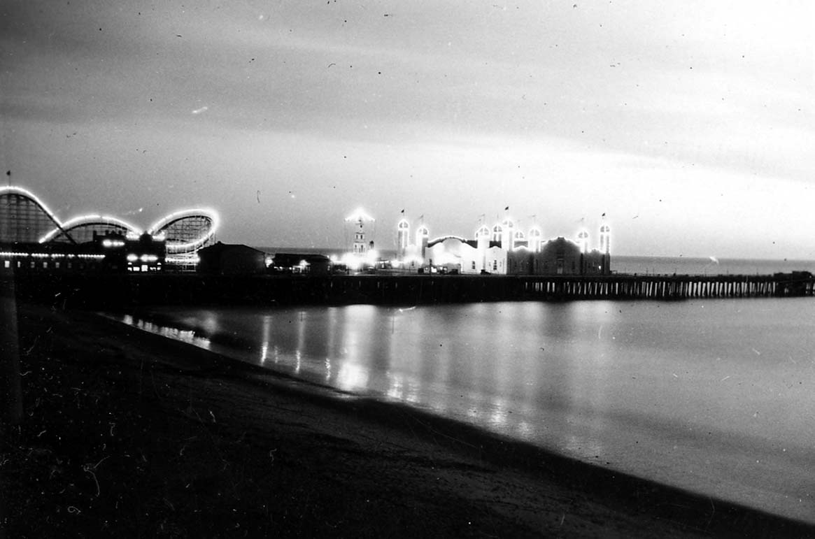 An evening view across the beach toward the illuminated Santa Monica Pier, showcasing the Moorish arches of the La Monica Ballroom and the adjacent roller coaster.