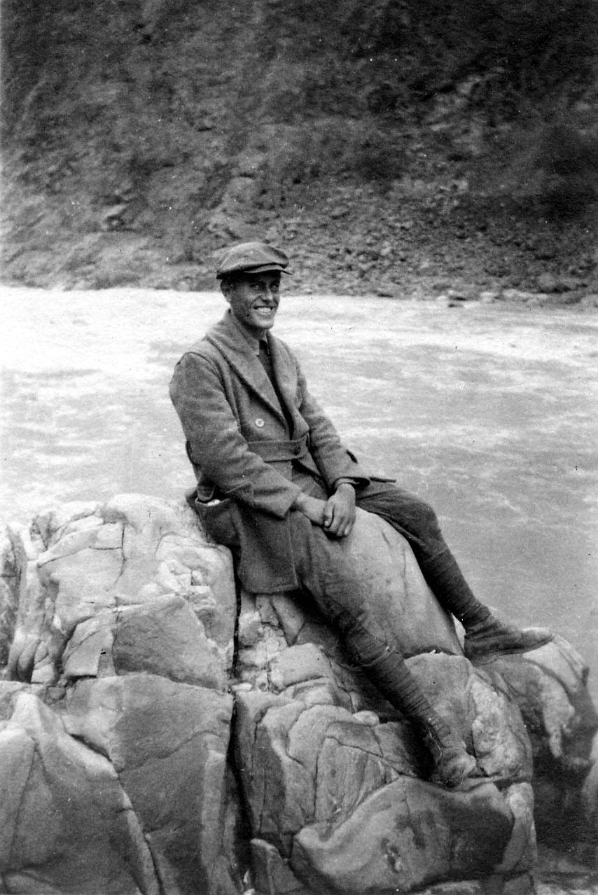 A smiling Donald A. Hall Sr. sitting on massive rocks beside a rushing river, wearing a heavy overcoat, tall boots, and a flat cap.