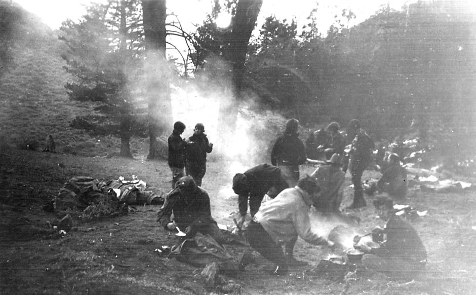 A large group of hikers gathered around a smoky campfire in a wooded clearing, cooking food in cast-iron pots.