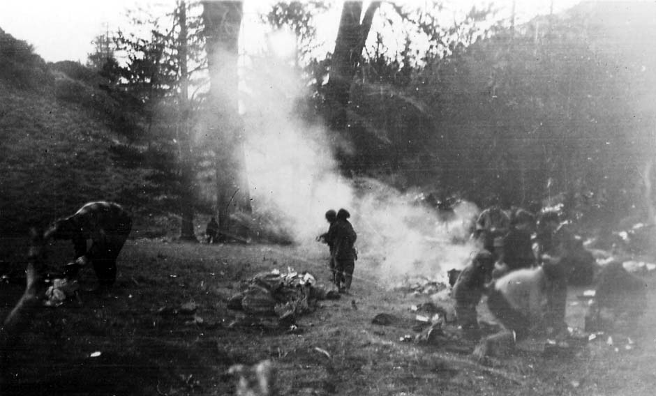 A group of hikers setting up camp in a wooded area, with heavy smoke billowing from a campfire as men unpack gear.