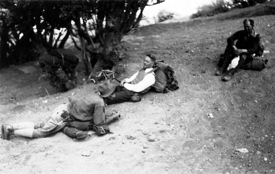 Three hikers with massive canvas backpacks sitting and reclining in the dirt on a steep hillside to recover.