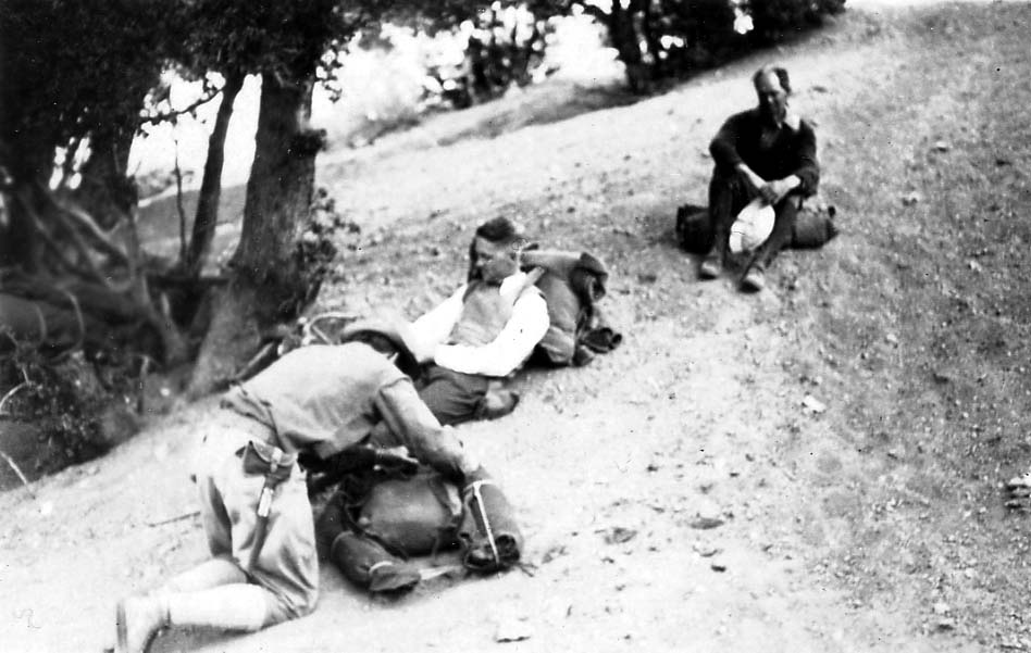 Three hikers with large, heavy backpacks sitting and lying in the dirt alongside a mountain trail to rest.