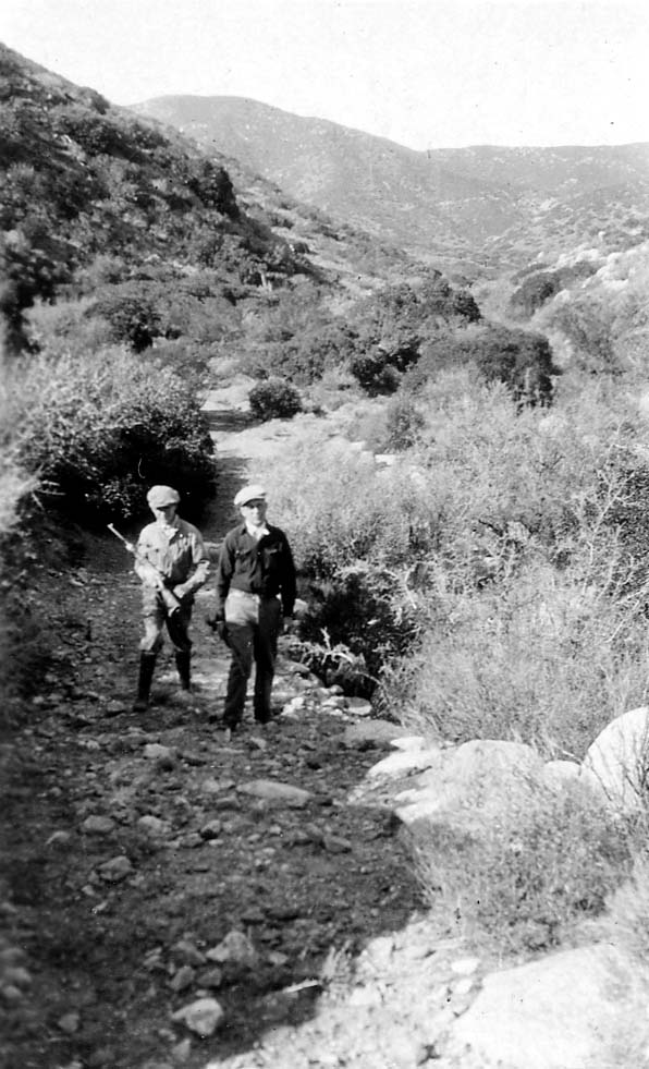 Two men wearing flat caps and hiking gear navigating a rocky, scrub-covered mountain trail.