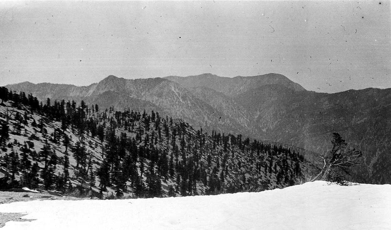 A wide landscape view of a rugged, snow-dusted mountain ridgeline stretching across the horizon under a cloudy sky.