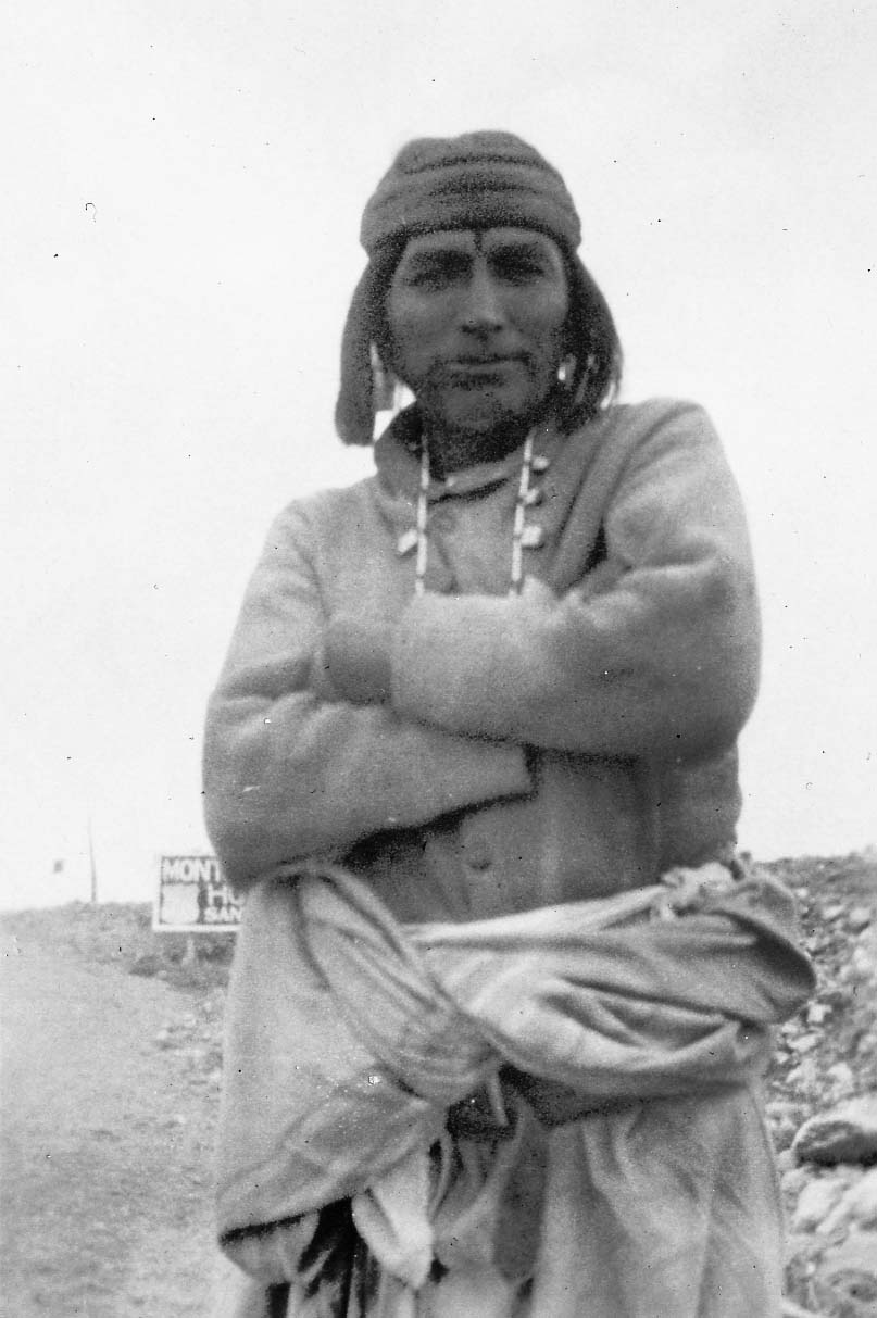 An outdoor portrait of a Southwest Indigenous man with folded arms, wearing a traditional headband, necklace, and blanket wrap beside a desert highway sign.