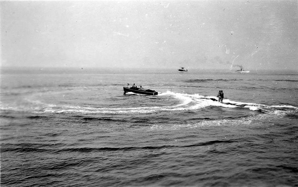 A wooden runabout speedboat carving a turn while towing an aquaplaner standing on a surf board, with a float biplane and ship in the distant background.