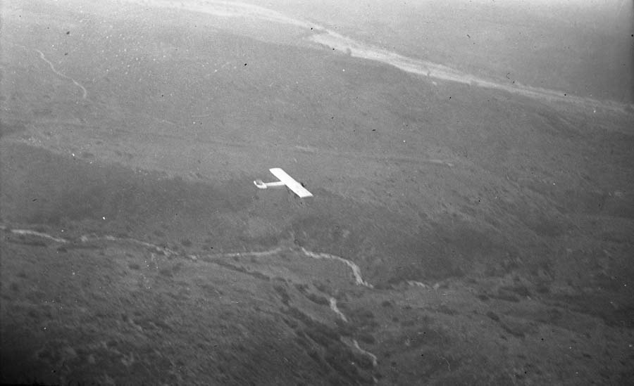 An air-to-air photograph taken from a chase plane showing a distant view of the fully completed Spirit of St. Louis flying over the California landscape during its departure.