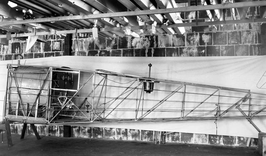 A wide view of the bare tubular steel fuselage of the Spirit of St. Louis, featuring the Earth Inductor Compass generator mounted on the upper frame behind the cockpit.