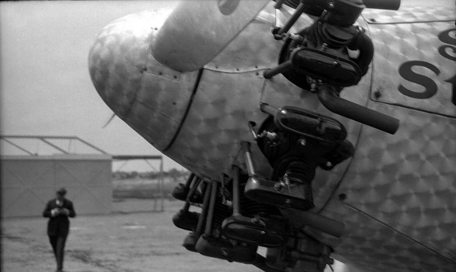 An artistic, low-angle shot focusing on the burnished engine cowling and cylinders of the Spirit of St. Louis, with Charles Lindbergh walking toward the aircraft in the background.