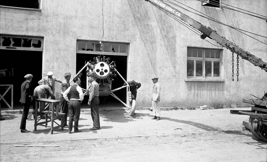 A crew of men, including Production Manager Hawley Bowlus, using a crane to lift the nose of the Spirit of St. Louis outside the factory doors after removing the landing gear.