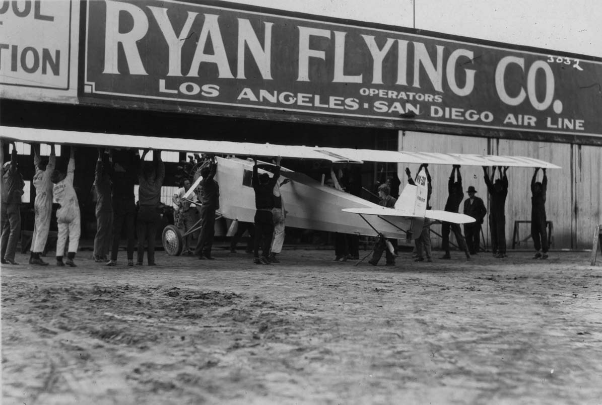 A large crew of men physically lifting the massive 46-foot wing of the Spirit of St. Louis to attach it to the fuselage outside the Ryan Flying Co. hangar.