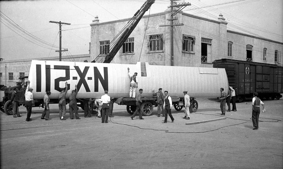 A large crew using a crane to carefully lower the massive Spirit of St. Louis wing from the train boxcar onto a waiting flatbed truck.