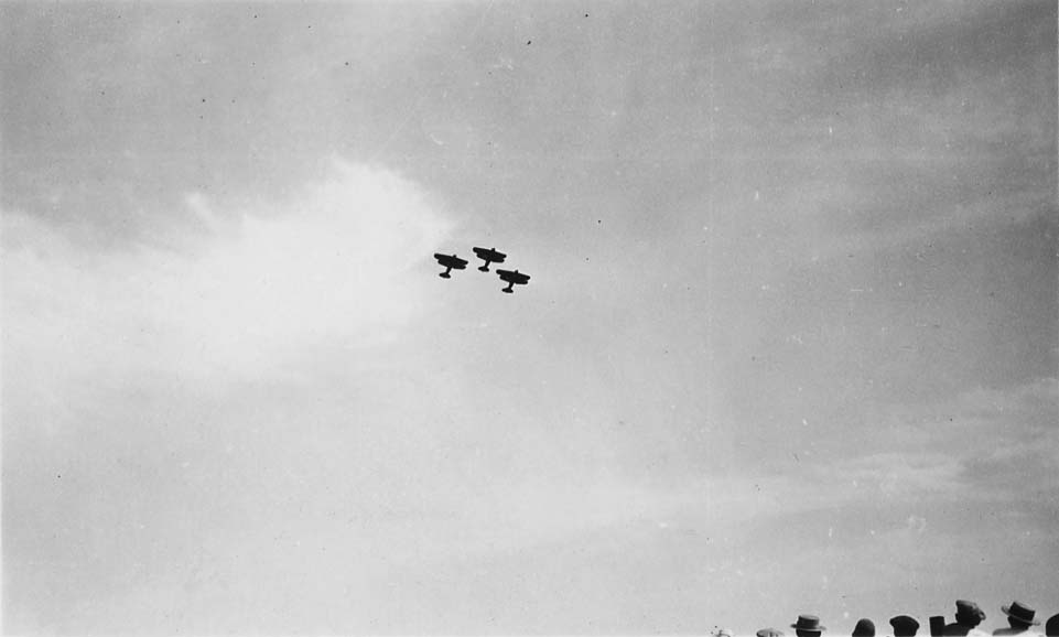 A very distant, high-altitude view of three biplanes flying in a tight V-formation against a bright sky.