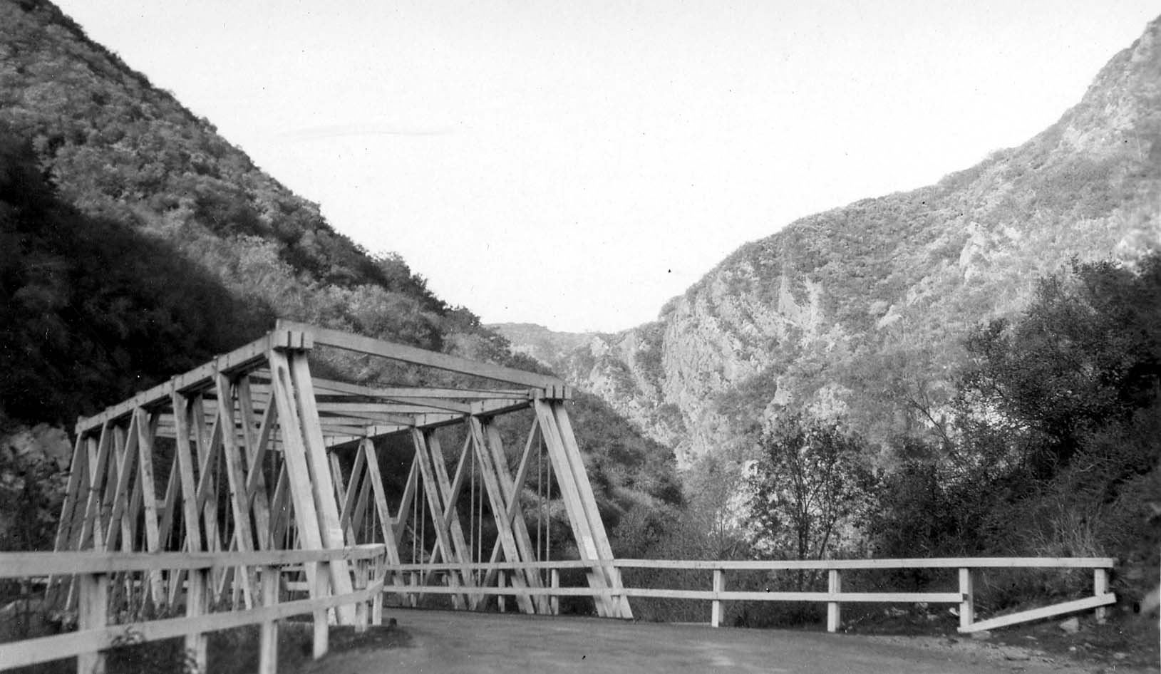 A heavy wooden truss road bridge crossing a creek in a rugged, steep mountain canyon flanked by brush-covered slopes.