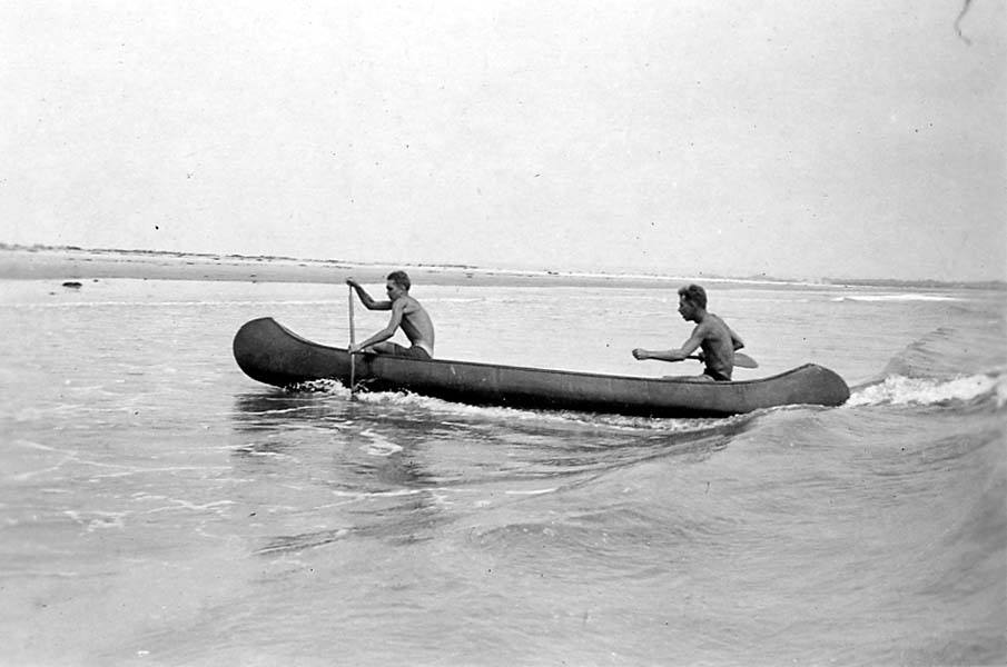 Two shirtless men paddling a canoe through the ocean surf, battling the white-capped waves near the shoreline.
