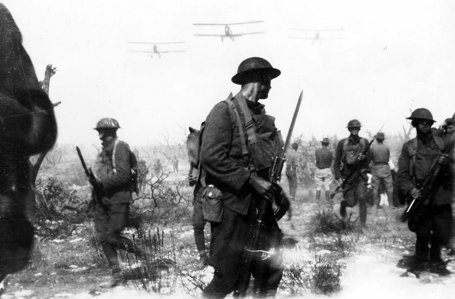 U.S. Army soldiers in Brodie helmets advancing with fixed bayonets across scrub terrain as three military biplanes fly overhead in V-formation.