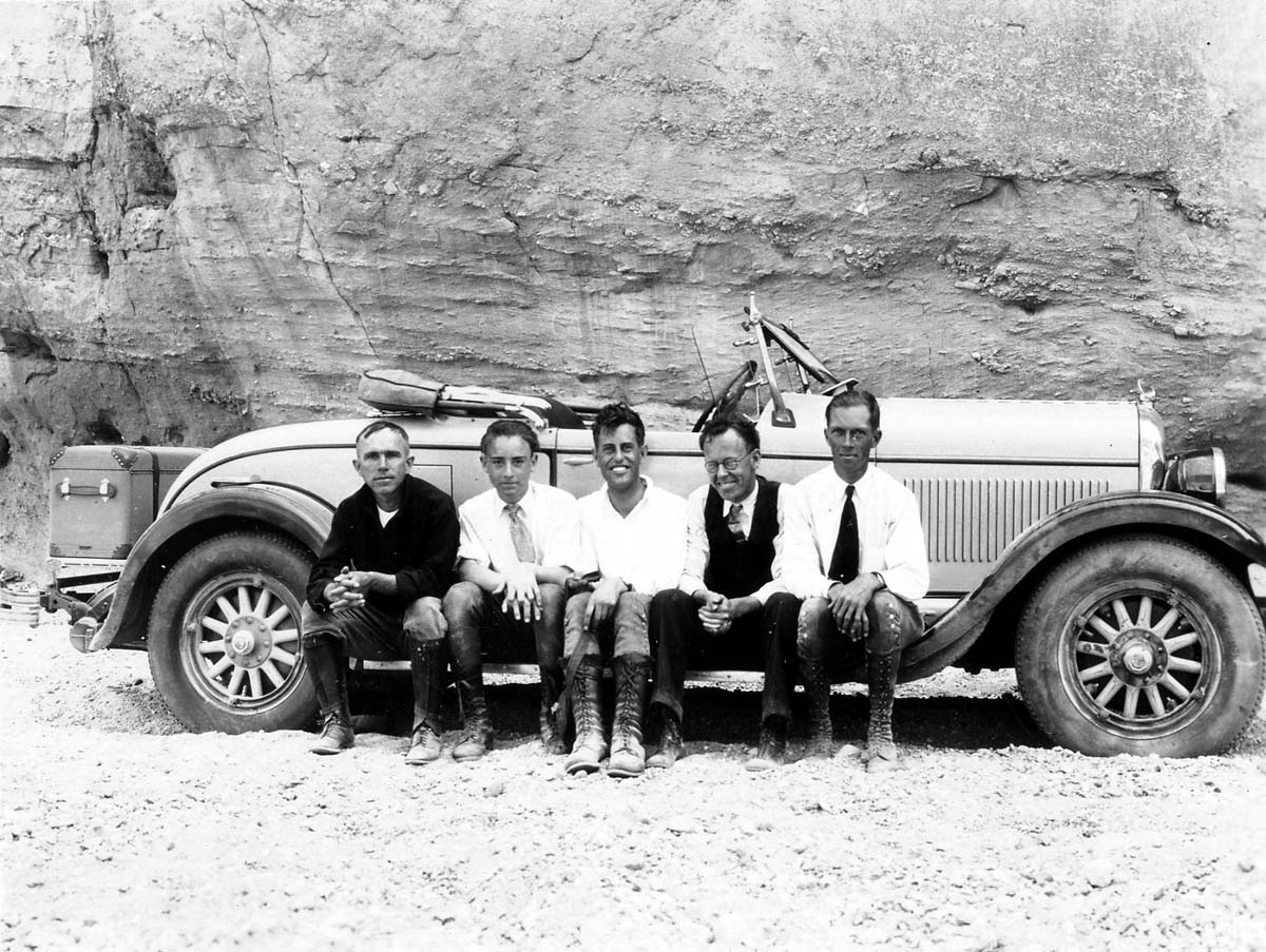 Five men sitting together on the running board of a vintage convertible automobile parked against a sheer rock wall.