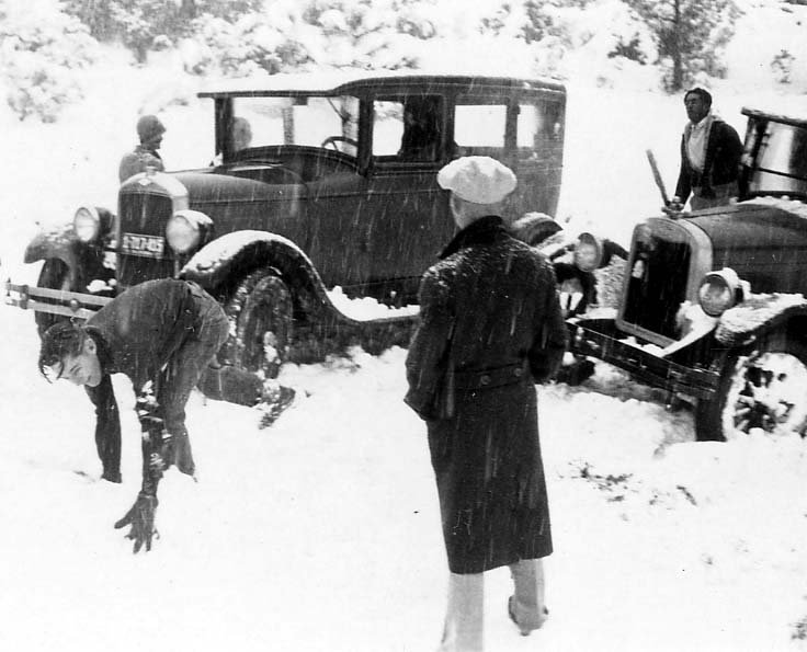 Three men attempting to dig out or push two vintage automobiles that are deeply stuck in heavy mountain snow.