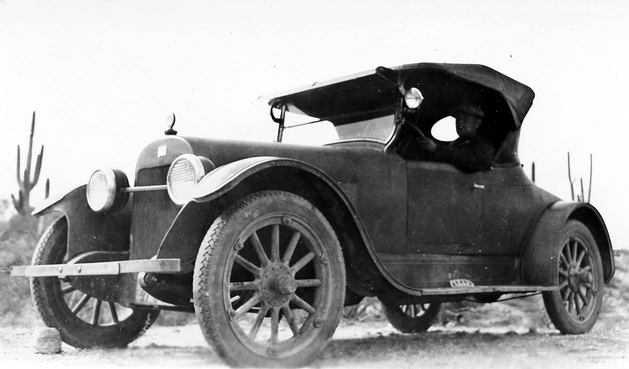 Donald Hall sitting in the driver's seat of an early vintage convertible automobile parked on a dirt road, framed against a stark sky with saguaro cactus in the background.