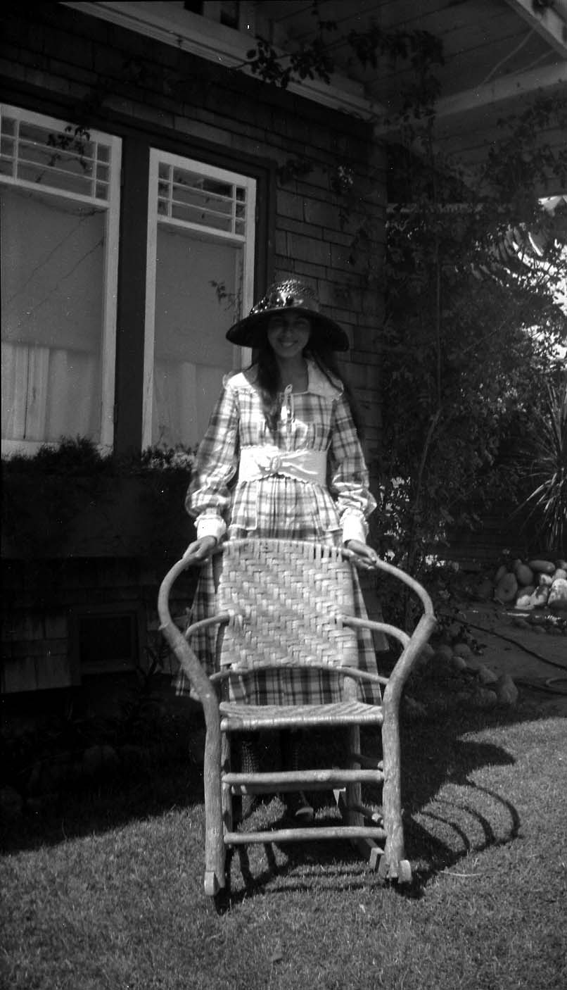 A young woman in a plaid dress and straw hat standing behind a woven wicker rocking chair in front of a Craftsman home.