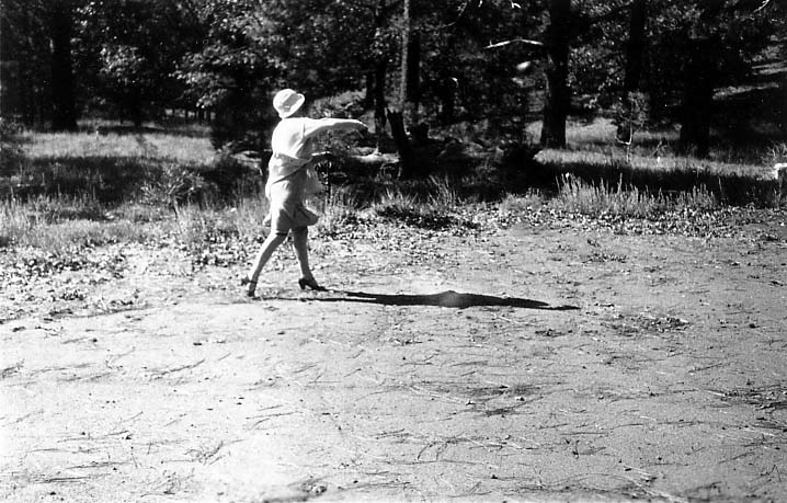 A woman wearing a 1920s dress, cloche hat, and high heels mid-throw with a baseball in a wooded clearing.