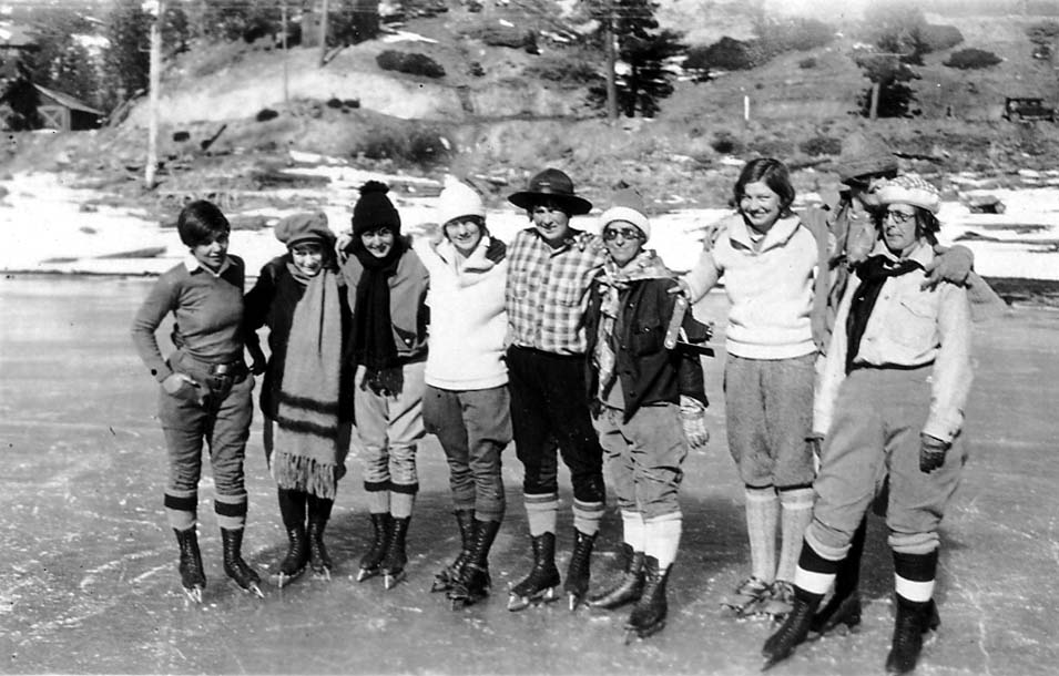 A group of eight women wearing winter clothing and ice skates, standing together in a line and smiling on a frozen lake.