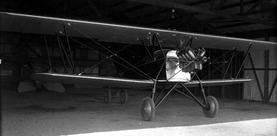 An open-cockpit biplane parked inside a dark corrugated metal hangar, showcasing an uncowled Wright-Hispano V-8 water-cooled engine.