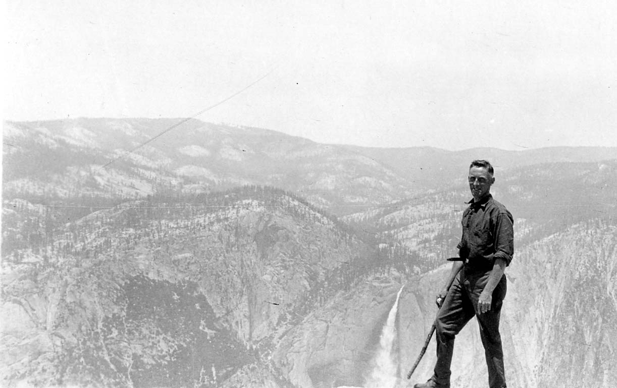 A hiker holding a walking stick standing on an exposed granite cliff ledge overlooking the massive canyon and plunge of Upper Yosemite Fall.