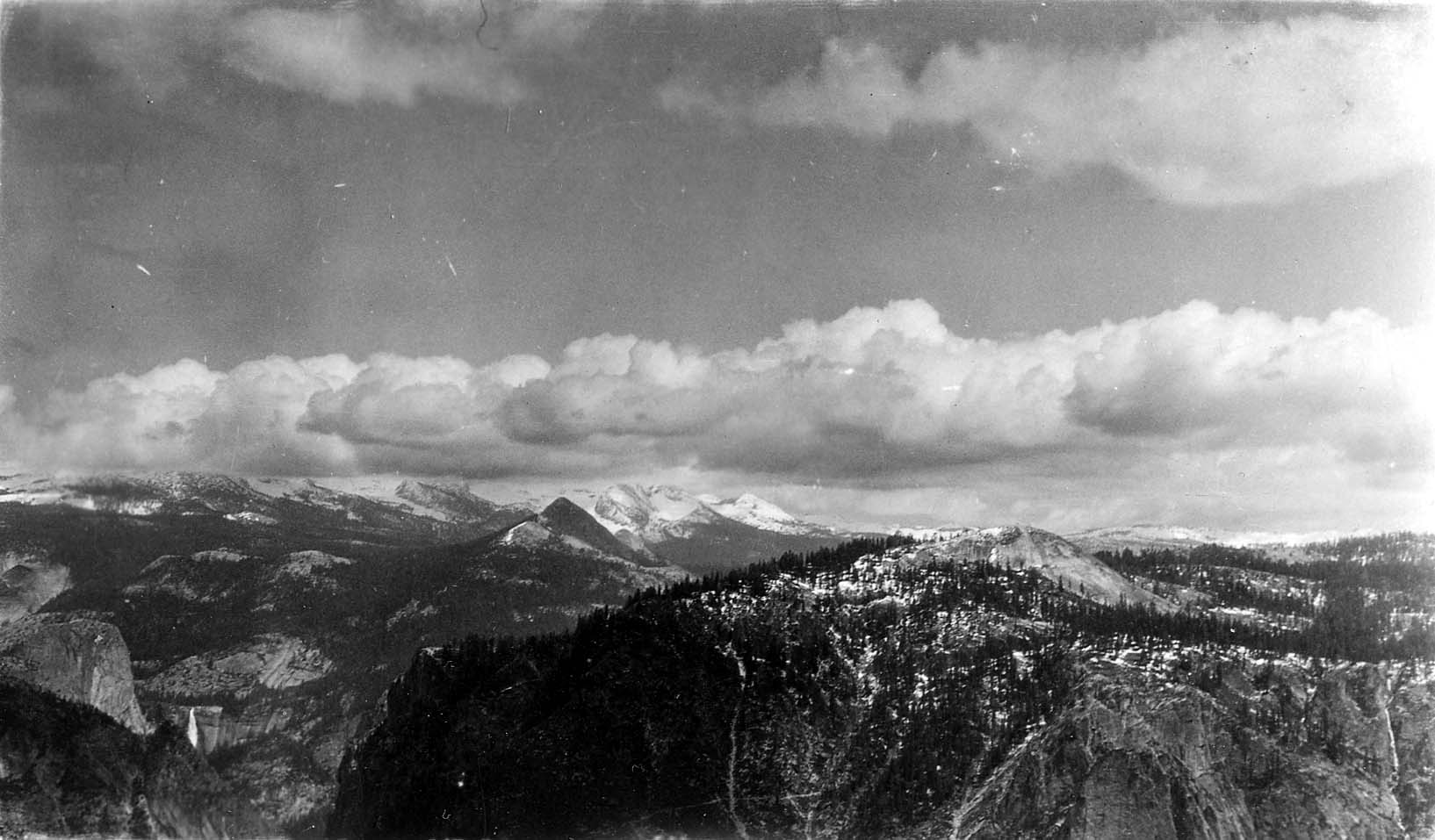 A sweeping panoramic vista from Glacier Point overlooking snow-dusted High Sierra peaks, granite domes, and valley canyons under cumulus clouds.