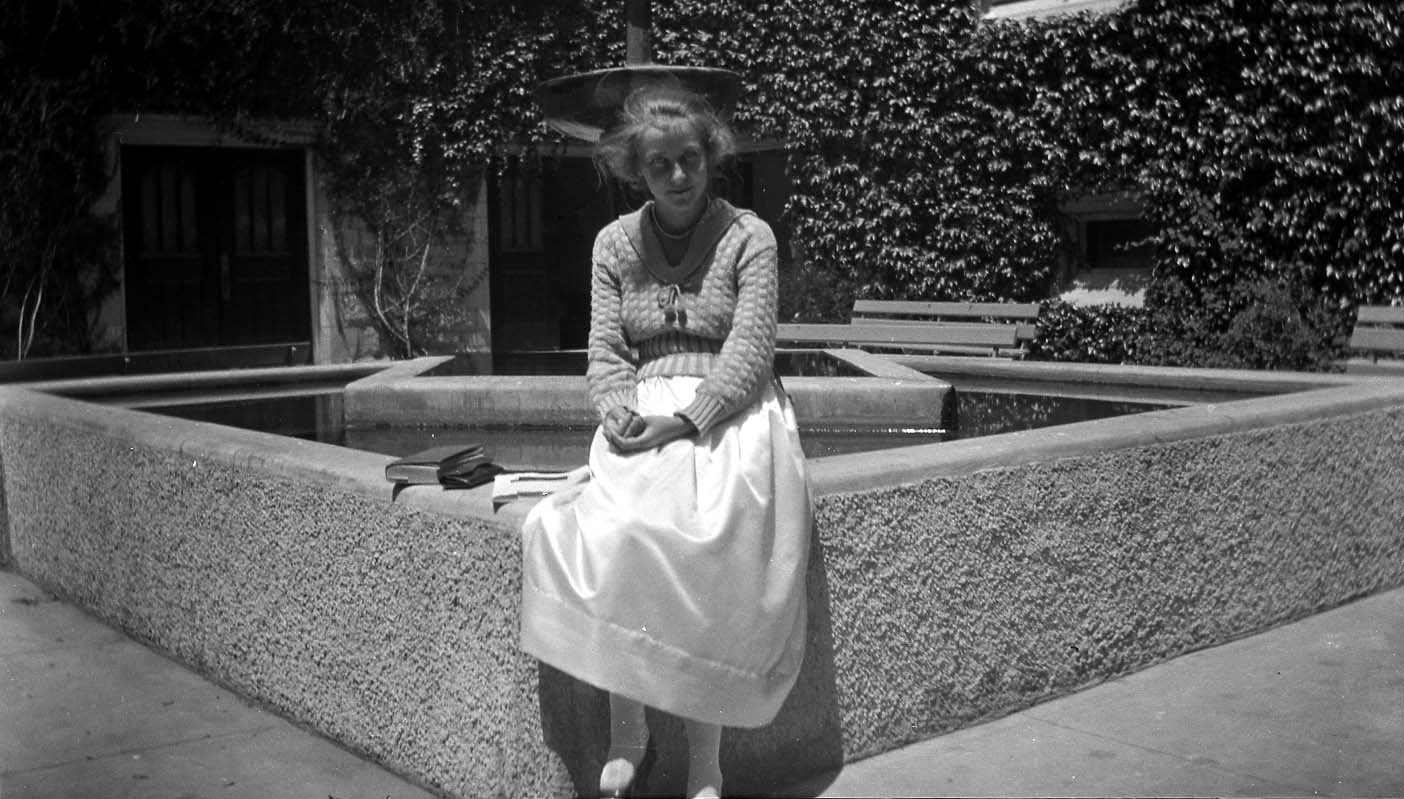 A young woman in a knitted sweater and white skirt seated on the concrete ledge of a fountain pool in an ivy-covered courtyard.