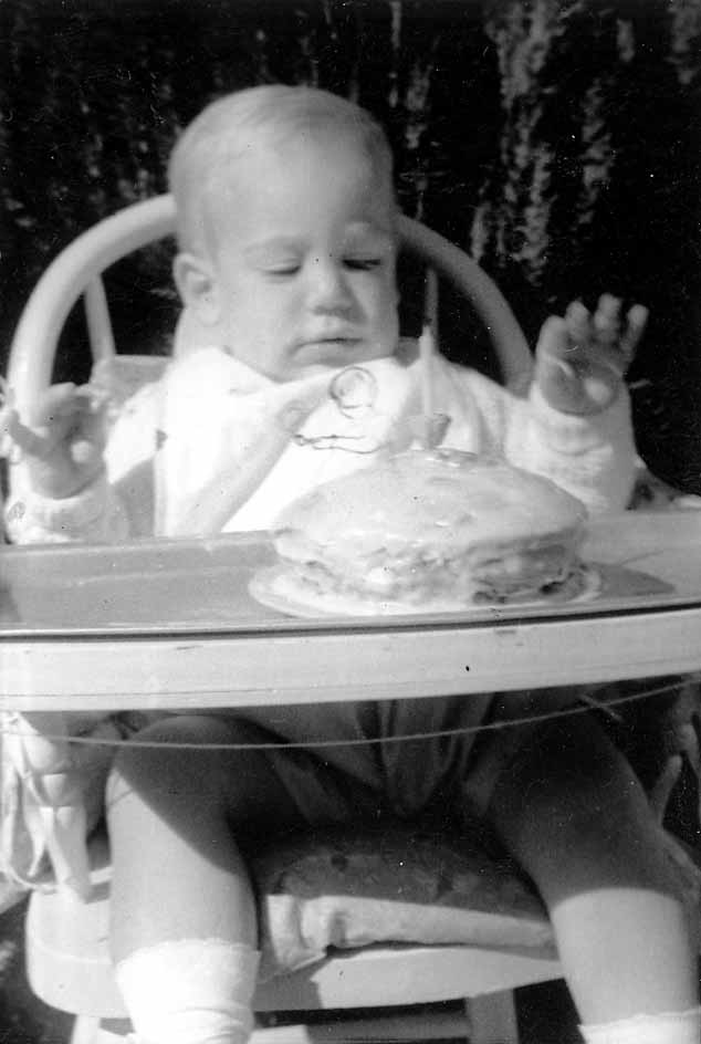 A toddler, Don Hall Jr., sitting in a highchair and looking down at a small birthday cake with a single candle on the tray.