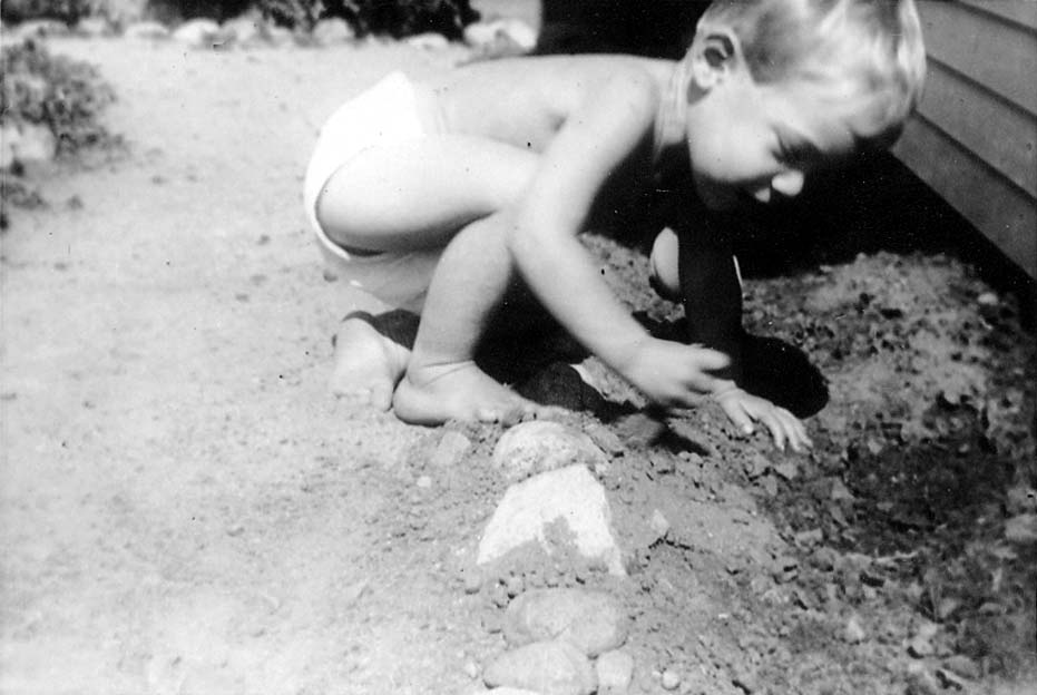 A young toddler, Don Hall Jr., crouching in the dirt next to a house foundation, playing with a line of rocks.