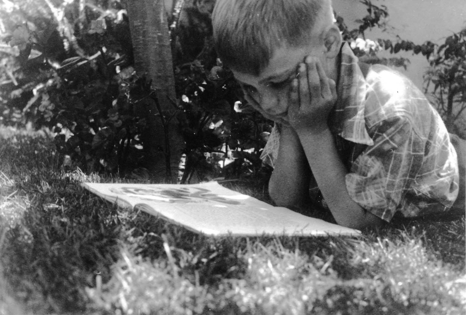 A young boy, Don Hall Jr., lying on his stomach in the grass, resting his head in his hands while looking down at an open book.
