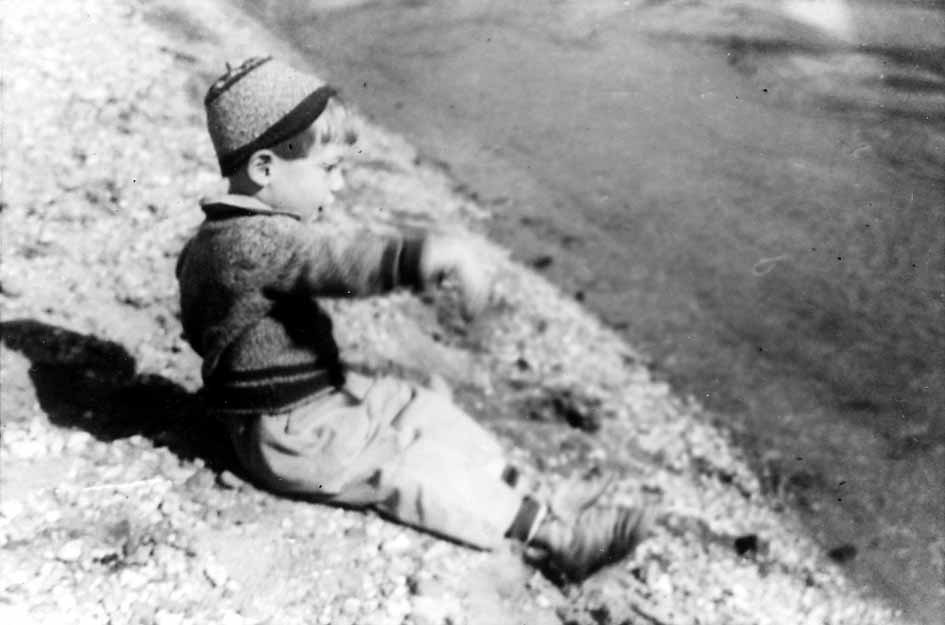 A young boy, Don Hall Jr., wearing a sweater and a small cap, sitting on a rocky beach and pointing out toward the water.