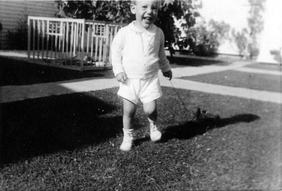 A toddler, Don Hall Jr., walking across a grassy yard in a white outfit, pulling a small toy on a string behind him.