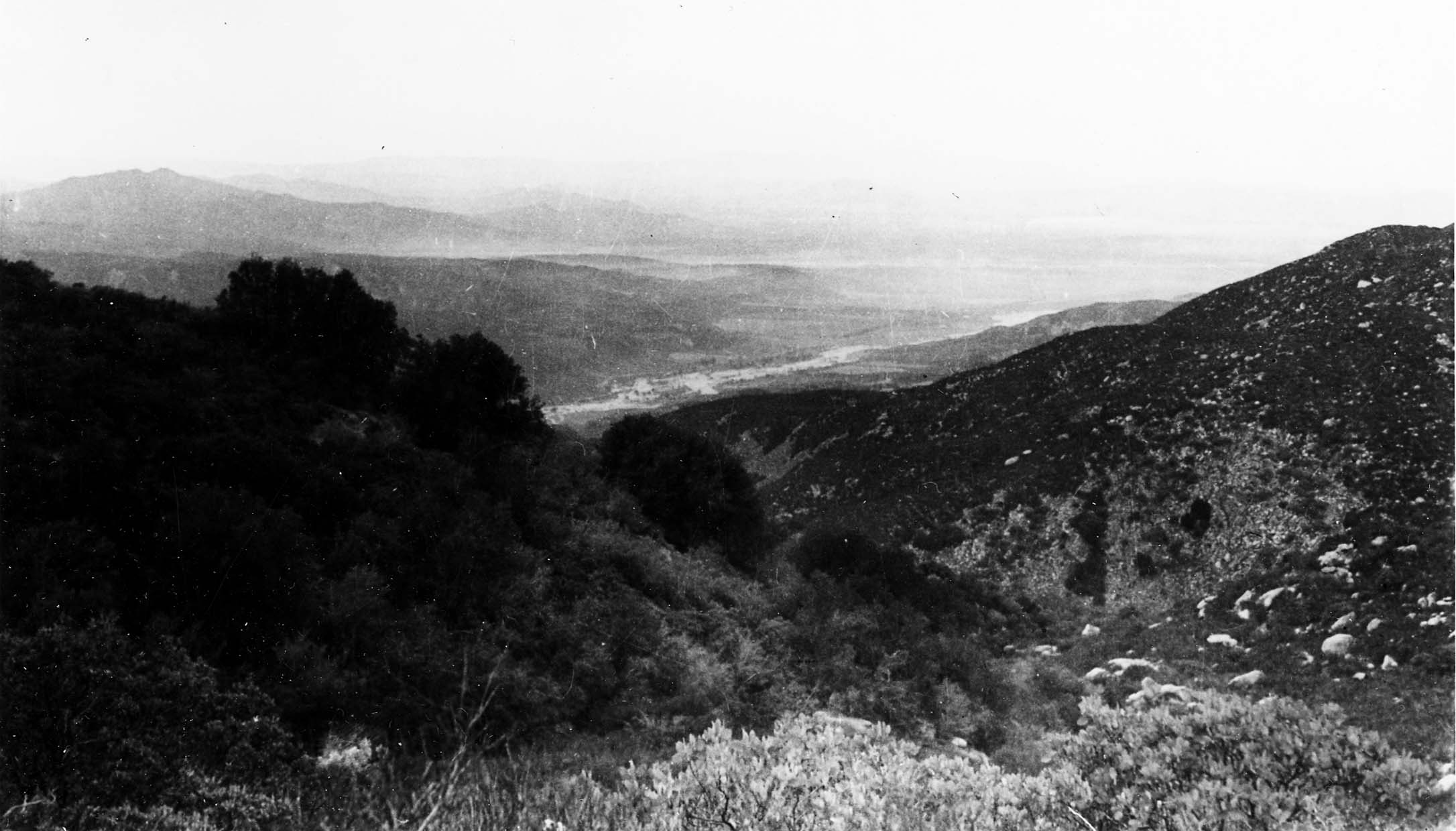 A high-altitude view looking down a heavily forested mountain valley toward a distant misty plain and river basin.