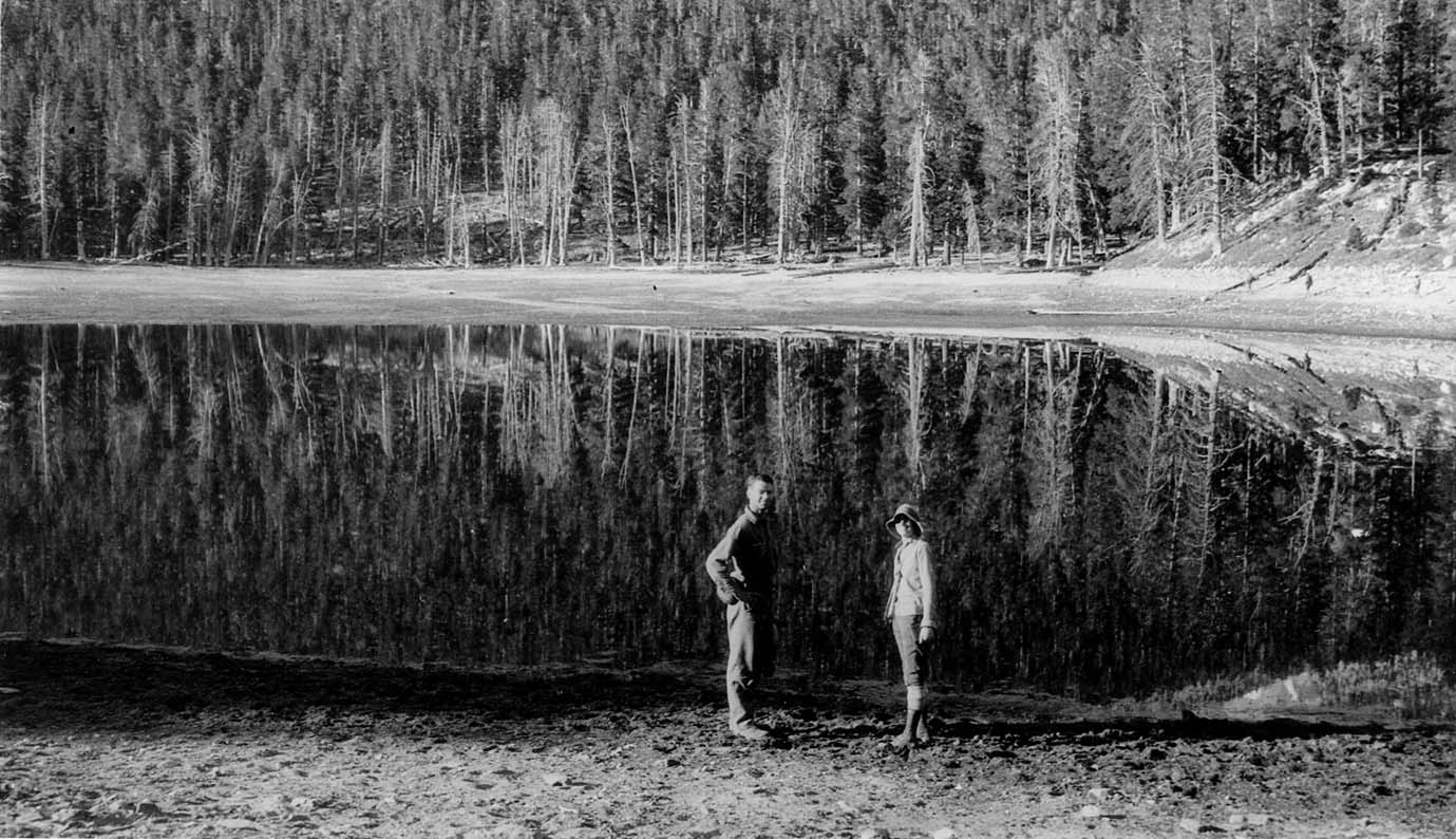 A glassy, mirror-like alpine lake reflecting a dense wall of tall evergreen trees along its shoreline, with two hikers standing on the bank.