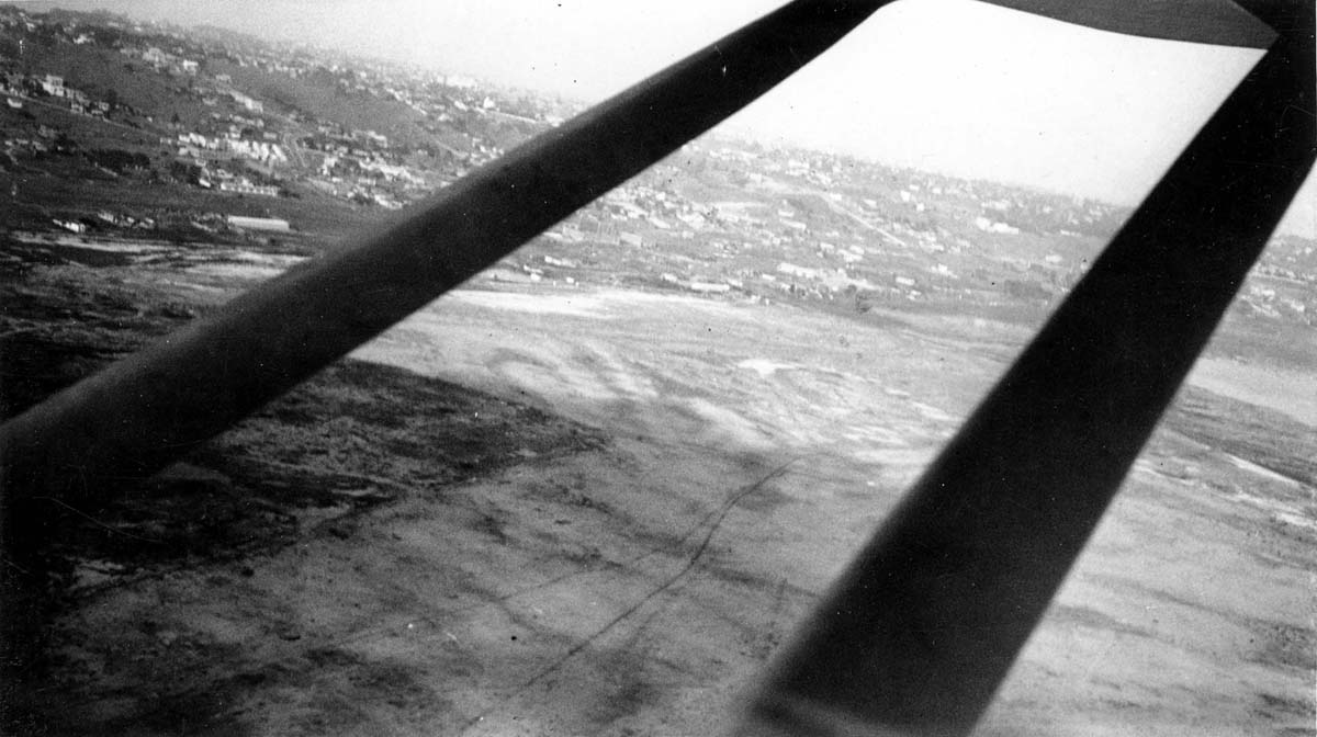 Aerial view between wing lift struts looking down at Dutch Flats dirt airfield and San Diego landscape.