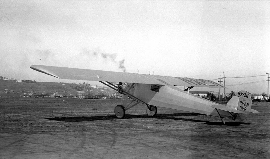 Left profile view of the Spirit of St. Louis showing rudder manufacturer markings and distant smoke plume over hills.